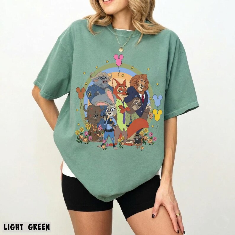 May include: Light green t-shirt featuring a colorful graphic of Zootopia characters. The design includes Judy Hopps, Nick Wilde, and other characters in a circular arrangement with flowers and balloons. The text "LIGHT GREEN" is at the bottom.