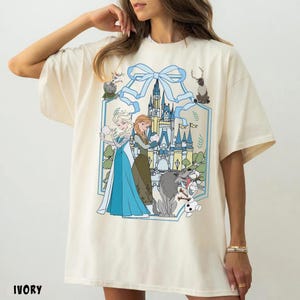 May include: Ivory-colored t-shirt featuring a graphic of two princesses, a castle, and various characters. The design is framed by a light blue bow and decorative elements. The word "IVORY" is printed at the bottom.