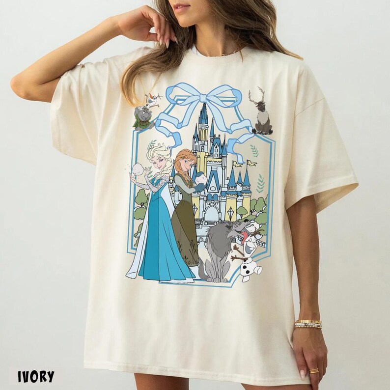 May include: Ivory-colored t-shirt featuring a graphic of two princesses, a castle, and various characters. The design is framed by a light blue bow and decorative elements. The word "IVORY" is printed at the bottom.