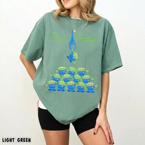 May include: Light green t-shirt featuring a graphic of the Pizza Planet logo and a claw crane with green aliens. The aliens are stacked in a pyramid shape. The text "LIGHT GREEN" is at the bottom.