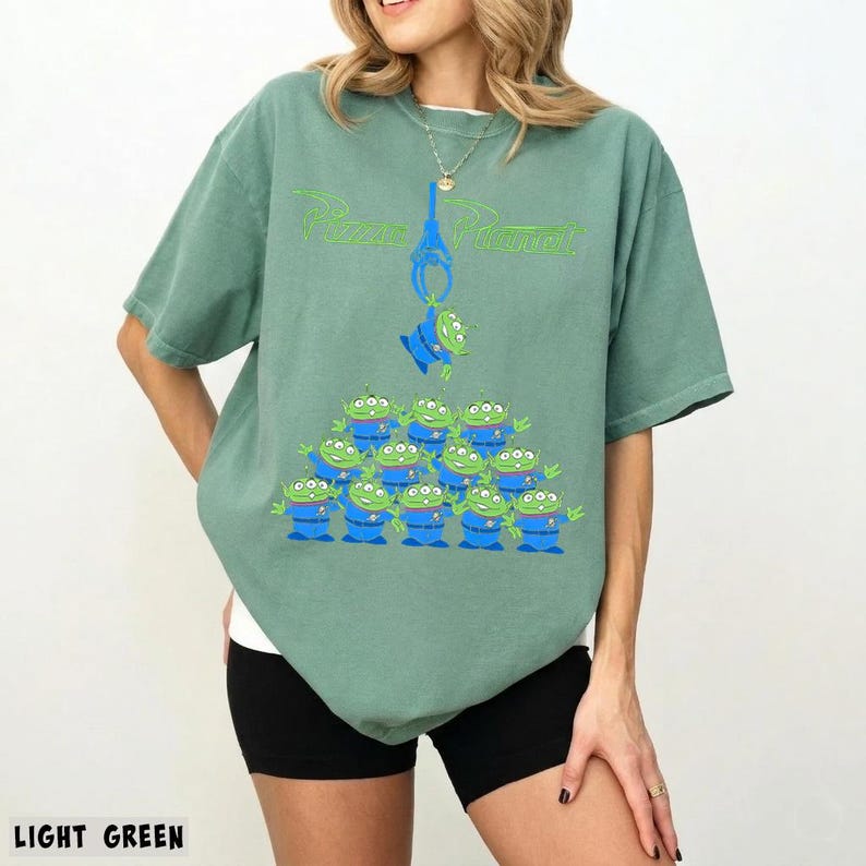 May include: Light green t-shirt featuring a graphic of the Pizza Planet logo and a claw crane with green aliens. The aliens are stacked in a pyramid shape. The text "LIGHT GREEN" is at the bottom.
