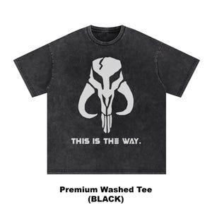 May include: Black washed tee featuring a white graphic of a skull-like helmet with the text "THIS IS THE WAY." The shirt is a premium washed tee.