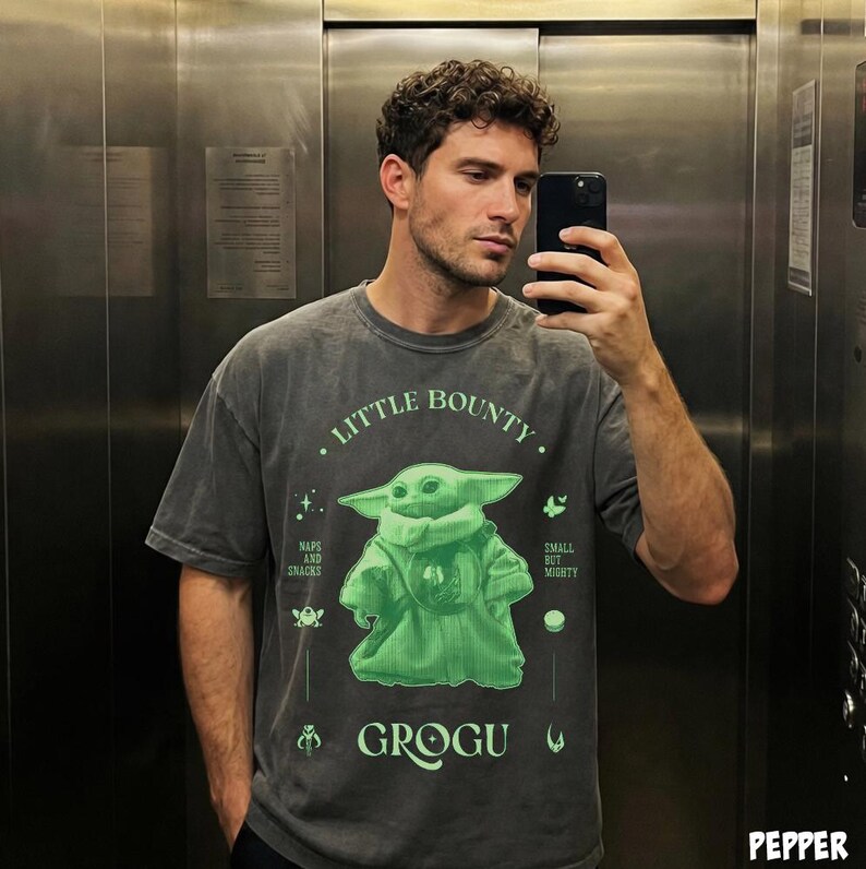 Op de afbeelding: Grijs t-shirt met een groene Grogu-afbeelding uit The Mandalorian. De tekst op het shirt luidt "Little Bounty", "Naps and Snacks", "Small but Mighty" en "Grogu".