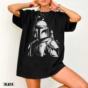 K&ouml;nnte beinhalten: Schwarzes T-Shirt mit einer Graustufen-Illustration einer Figur mit Helm. Das Design ist zentriert, und das Wort "BLACK" ist unten aufgedruckt. Die Grafik ist ein detailliertes, stilisiertes Portr&auml;t.