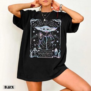 May include: Black t-shirt featuring a graphic of Grogu from Star Wars. The design includes the text "Grogu" and "Star Wars" in a stylized font, with a colorful, holographic effect. The t-shirt is a classic crew neck style.