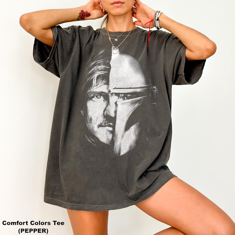 May include: Pepper-colored Comfort Colors tee featuring a split image of a man's face and a Mandalorian helmet. The graphic is in black and white. The shirt has short sleeves and a relaxed fit.