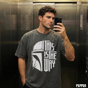 May include: Dark grey t-shirt with a white helmet graphic and the words "THIS IS THE WAY." The shirt has a faded appearance. The design is centrally placed on the front of the garment.