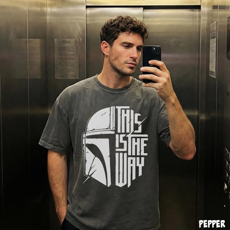 May include: Dark grey t-shirt with a white helmet graphic and the words "THIS IS THE WAY." The shirt has a faded appearance. The design is centrally placed on the front of the garment.