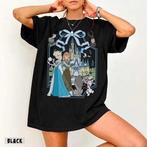 May include: Black t-shirt featuring a cartoon illustration of Elsa and Anna from Frozen, with a castle and other characters. The design is framed by a blue ribbon and a hexagonal border. The word "BLACK" is printed at the bottom.