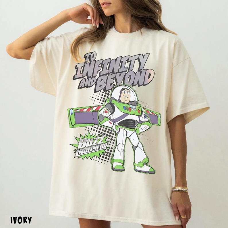 May include: An ivory t-shirt featuring a graphic of the character Buzz Lightyear with the text "TO INFINITY AND BEYOND" and "BUZZ LIGHTYEAR" in a bold, stylized font. The design includes a comic-book style dot pattern and green accents.