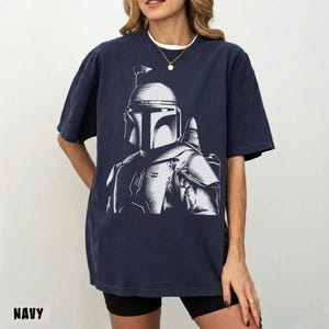 K&ouml;nnte beinhalten: Marineblaues T-Shirt mit einer wei&szlig;en Grafik einer Science-Fiction-Figur. Die Grafik ist eine detaillierte Illustration einer Figur mit Helm. Das Wort "NAVY" ist unten auf dem T-Shirt aufgedruckt.