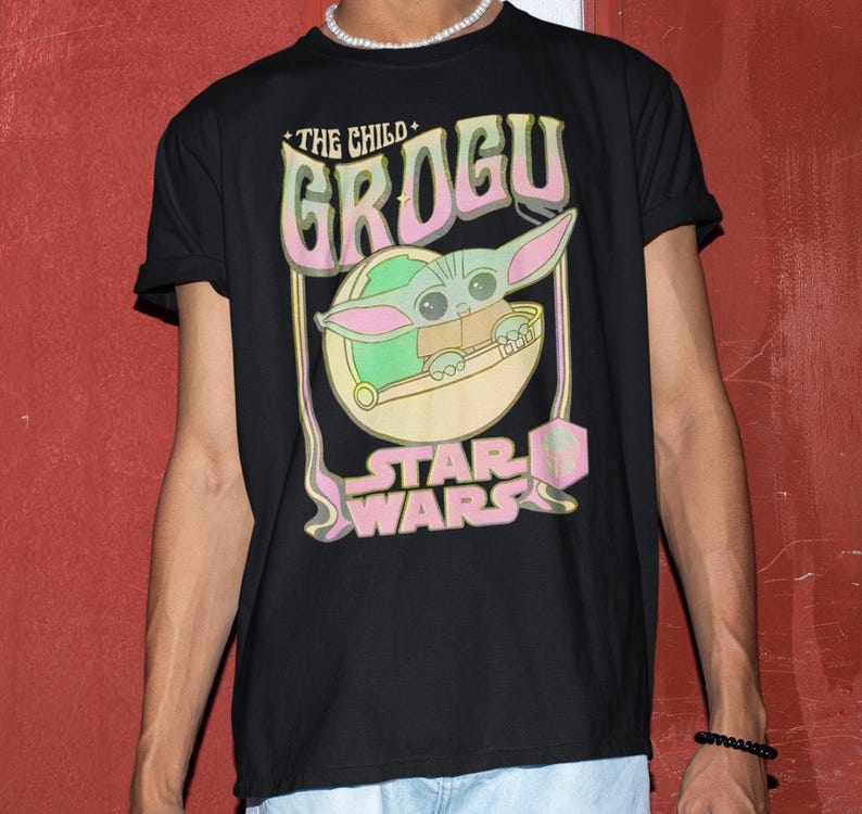 May include: Black t-shirt featuring a Grogu graphic from Star Wars. The design includes the text "THE CHILD GROGU" in pink and green, with Grogu in a pod. The Star Wars logo is at the bottom.