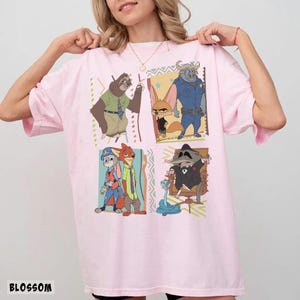 Puede incluir: Una camiseta rosa claro con personajes de dibujos animados de Zootr&oacute;polis. La camiseta tiene cuatro paneles con diferentes personajes, incluyendo un perezoso, un zorro y un oficial de polic&iacute;a. La palabra "BLOSSOM" est&aacute; impresa en la parte inferior izquierda.