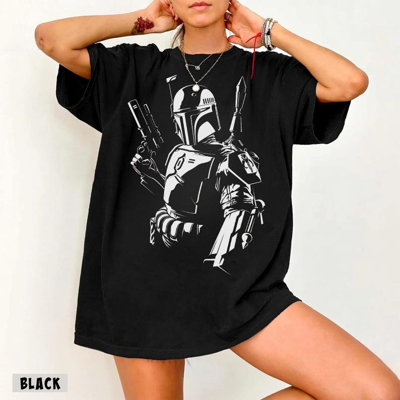 May include: Black t-shirt featuring a white graphic of a bounty hunter with a helmet, holding a blaster and a spear. The word "BLACK" is printed at the bottom of the shirt. The shirt has short sleeves and a relaxed fit.