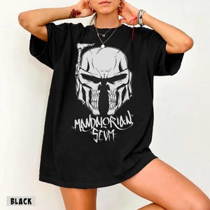 Pu&ograve; includere: T-shirt nero con grafica bianca di un elmo a forma di teschio e la scritta "Mandalorian Scum" in un font stilizzato. La maglietta &egrave; di colore nero pieno.