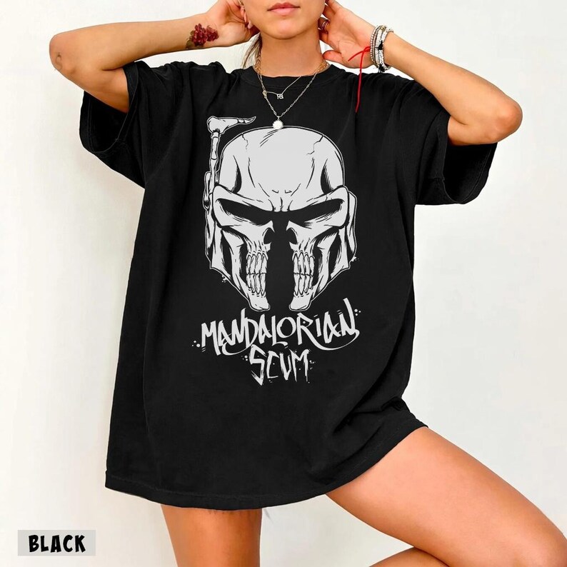 Pu&ograve; includere: T-shirt nero con grafica bianca di un elmo a forma di teschio e la scritta "Mandalorian Scum" in un font stilizzato. La maglietta &egrave; di colore nero pieno.