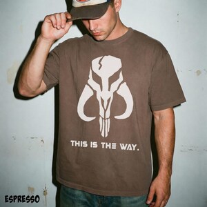 May include: Espresso-colored t-shirt featuring a white graphic of a skull with curved horns and the text "THIS IS THE WAY." The shirt is a short-sleeved crew neck.
