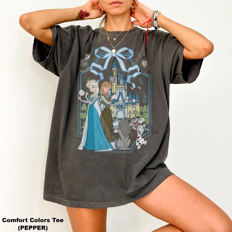 May include: Dark grey Comfort Colors tee featuring a graphic of Elsa and Anna from Frozen, with a castle backdrop and characters like Olaf and Sven. The design is framed by a blue ribbon and floral elements.