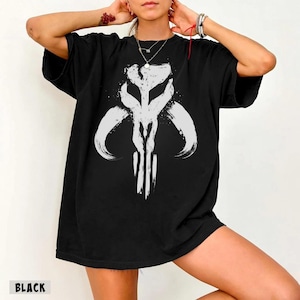 May include: Black t-shirt featuring a white graphic design. The design is a stylized, symmetrical emblem with curved and vertical lines. The word "BLACK" is printed at the bottom of the shirt.