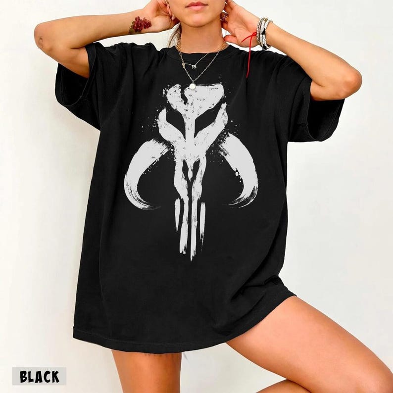 May include: Black t-shirt featuring a white graphic design. The design is a stylized, symmetrical emblem with curved and vertical lines. The word "BLACK" is printed at the bottom of the shirt.