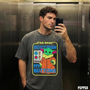 May include: Dark gray t-shirt featuring a graphic of Grogu from Star Wars with the text "DON'T PUSH MY BUTTONS". The design includes a green Grogu wearing an orange coat. The shirt has a boxy fit and short sleeves.