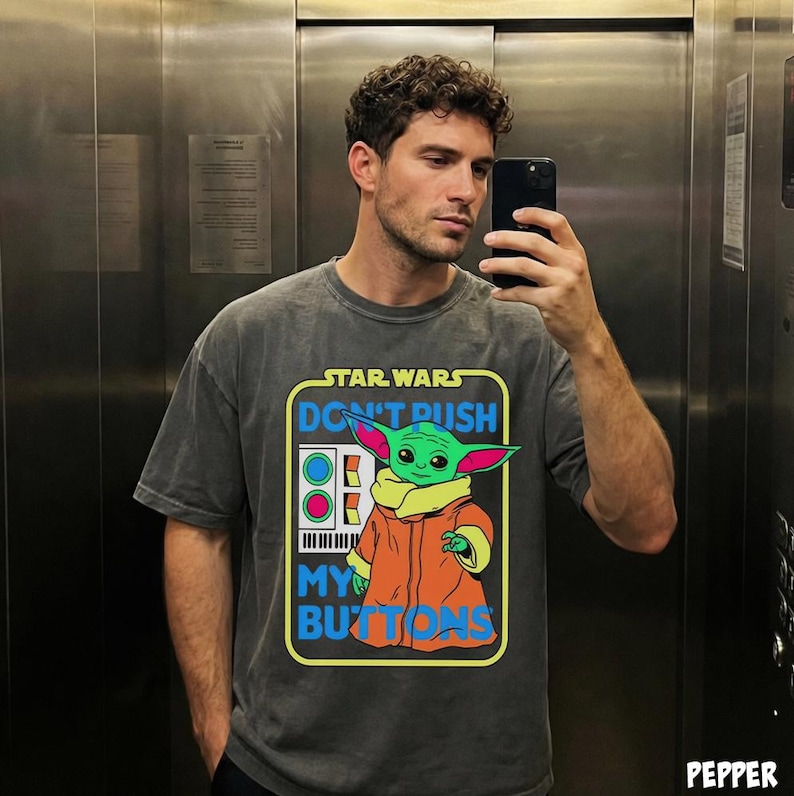 May include: Dark gray t-shirt featuring a graphic of Grogu from Star Wars with the text "DON'T PUSH MY BUTTONS". The design includes a green Grogu wearing an orange coat. The shirt has a boxy fit and short sleeves.