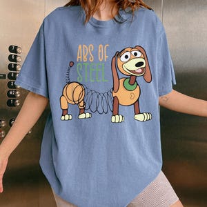 May include: A blue short-sleeve t-shirt featuring a graphic of a brown dachshund toy with a metal spring body. The text "ABS OF STEEL" is printed in orange and green above the character. The shirt has a crew neckline and a relaxed fit.