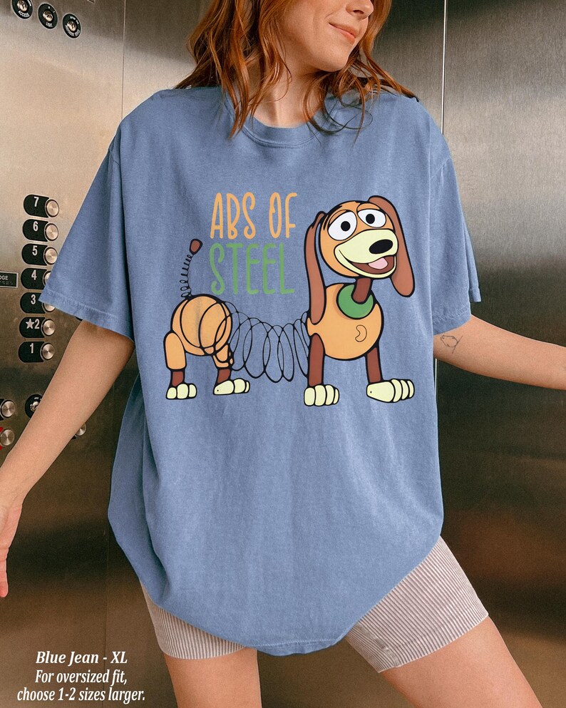 May include: A blue short-sleeve t-shirt featuring a graphic of a brown dachshund toy with a metal spring body. The text "ABS OF STEEL" is printed in orange and green above the character. The shirt has a crew neckline and a relaxed fit.