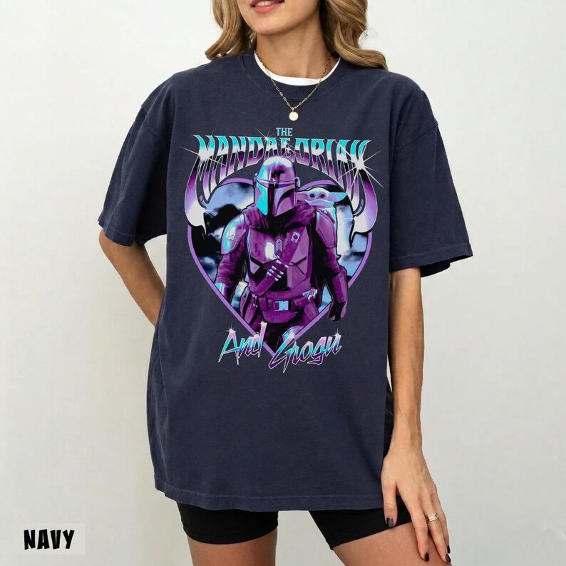 May include: Navy blue t-shirt featuring a graphic of The Mandalorian and Grogu. The design includes the text "The Mandalorian" in a stylized font above the image of the character. The text "And Grogu" is below the image. The word "NAVY" is at the bottom left.