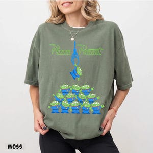 May include: Moss green t-shirt featuring a graphic of the Pizza Planet logo and a group of green alien characters. The aliens are wearing blue uniforms. The text "Pizza Planet" is in green.