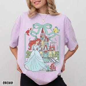 May include: Orchid-colored t-shirt featuring a cartoon illustration of a princess in a blue gown, a castle, and sea creatures. The design is framed by a green ribbon and a hexagonal border. The word "ORCHID" is printed at the bottom.