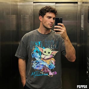 May include: A grey t-shirt with a graphic of Grogu from The Mandalorian and the Star Wars logo. The design includes a blue and orange spaceship and the text "Star Wars" and "The Mandalorian".