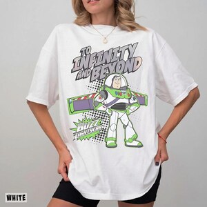 May include: A white short-sleeve t-shirt featuring a graphic of Buzz Lightyear with the text "TO INFINITY AND BEYOND" and "BUZZ LIGHTYEAR" in a comic book style. The design includes green, purple, and black accents on a white cotton fabric base.