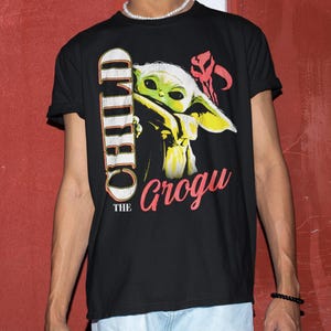 May include: Black t-shirt featuring a graphic of Grogu, also known as The Child, from The Mandalorian. The design includes the text "CHILD" vertically and "Grogu" in pink, with a red symbol. The shirt is a crew neck.