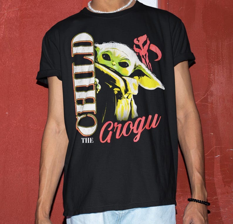 May include: Black t-shirt featuring a graphic of Grogu, also known as The Child, from The Mandalorian. The design includes the text "CHILD" vertically and "Grogu" in pink, with a red symbol. The shirt is a crew neck.
