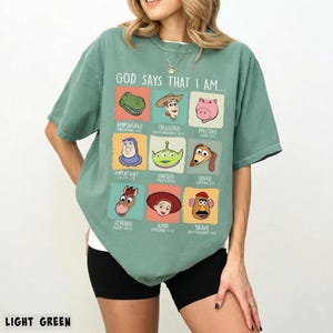 May include: Light green t-shirt with the text "GOD SAYS THAT I AM..." and cartoon character graphics. The shirt features a crew neck and short sleeves. The bottom of the shirt is visible.