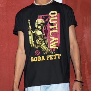Puede incluir: Camiseta negra con un gr&aacute;fico de Boba Fett, personaje de Star Wars, y la palabra "OUTLAW" en rosa y amarillo. El dise&ntilde;o tiene un aspecto vintage y desgastado.