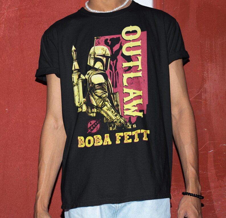 Puede incluir: Camiseta negra con un gr&aacute;fico de Boba Fett, personaje de Star Wars, y la palabra "OUTLAW" en rosa y amarillo. El dise&ntilde;o tiene un aspecto vintage y desgastado.