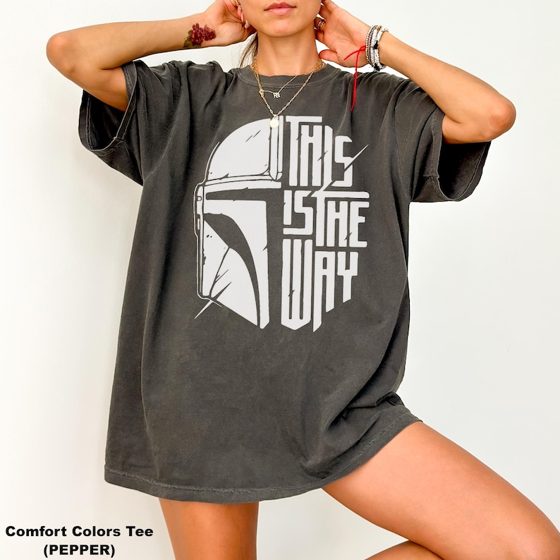May include: Dark grey Comfort Colors tee with a white graphic of a helmet and the text "THIS IS THE WAY." The shirt has short sleeves and a relaxed fit. The text is in a bold, stylized font.