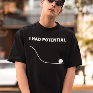May include: Black t-shirt with the white text "I HAD POTENTIAL" and a white graphic of a ball rolling down a curved line. The shirt is short-sleeved and worn by a person wearing a black cap and sunglasses.