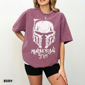 Pu&ograve; includere: T-shirt color bacca con grafica bianca di un casco a forma di teschio e la scritta "Mandalorian Scum". La maglietta ha girocollo e maniche corte. La parola "BERRY" &egrave; stampata in basso a sinistra.