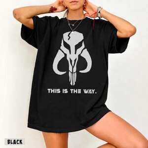 May include: Black t-shirt featuring a white graphic of a skull-like design with curved horns and vertical lines. The text "THIS IS THE WAY" is printed below the graphic. The word "BLACK" is printed at the bottom.