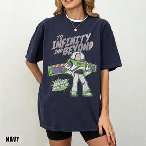 May include: A navy blue short-sleeve t-shirt featuring a graphic of the character Buzz Lightyear. The text "TO INFINITY AND BEYOND" is printed in a light gray font above the character, with the name "BUZZ LIGHTYEAR" in a green starburst below.