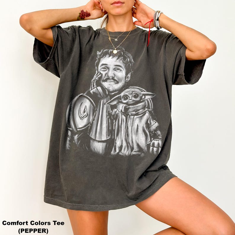 Puede incluir: Camiseta Comfort Colors color pimienta con un gr&aacute;fico en blanco y negro de un hombre y una peque&ntilde;a criatura. El hombre lleva una armadura y la criatura est&aacute; envuelta en una capa. El texto "Comfort Colors Tee (PEPPER)" est&aacute; en la parte inferior.