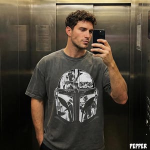 May include: A grey t-shirt with a black and white graphic of a helmet and character from a science fiction series. The shirt has a crew neck and short sleeves. The person in the image is wearing the shirt.