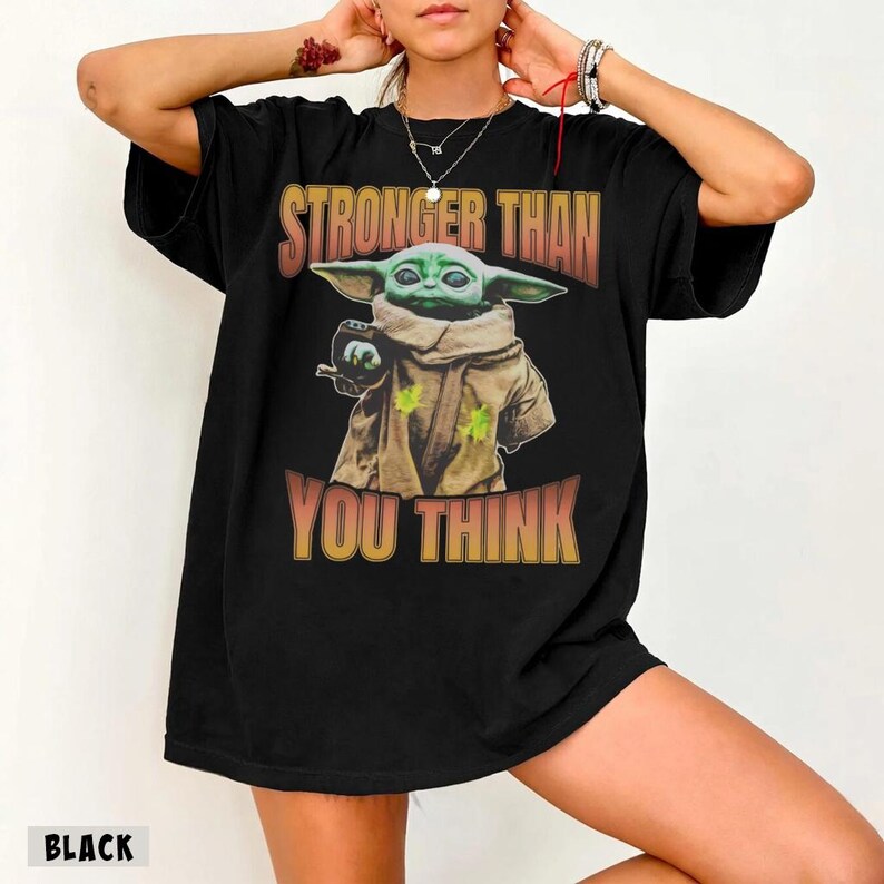 Pode incluir: Camiseta preta com uma estampa de Grogu, tamb&eacute;m conhecido como Baby Yoda, de The Mandalorian. O design inclui o texto "STRONGER THAN YOU THINK" em laranja e amarelo. A camiseta tem um corte folgado.