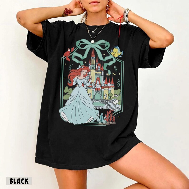 May include: Black t-shirt featuring a colorful illustration of a princess in a light blue gown, standing in front of a castle. The design includes a large bow, a crab, a fish, and the word "BLACK" at the bottom.