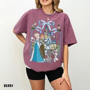 May include: Berry-colored t-shirt featuring a graphic of Elsa and Anna from Frozen, with a castle and other characters. The design is framed by a blue bow. The word "BERRY" is printed at the bottom of the shirt.