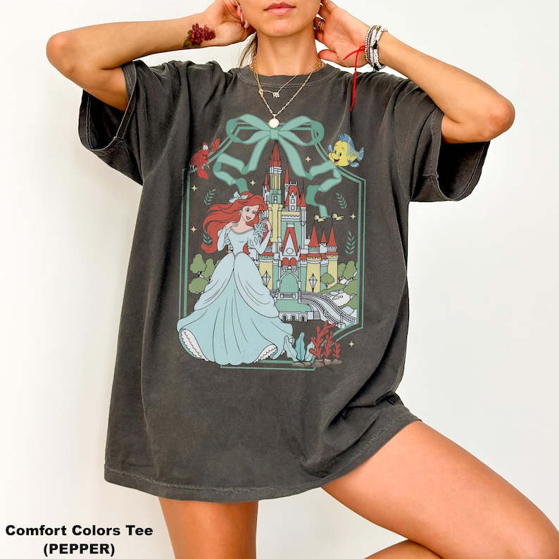 May include: A dark grey Comfort Colors tee featuring a graphic of a princess in a light blue gown, standing in front of a castle. The design includes a green bow, a fish, and other decorative elements. The text "Comfort Colors Tee (PEPPER)" is at the bottom.