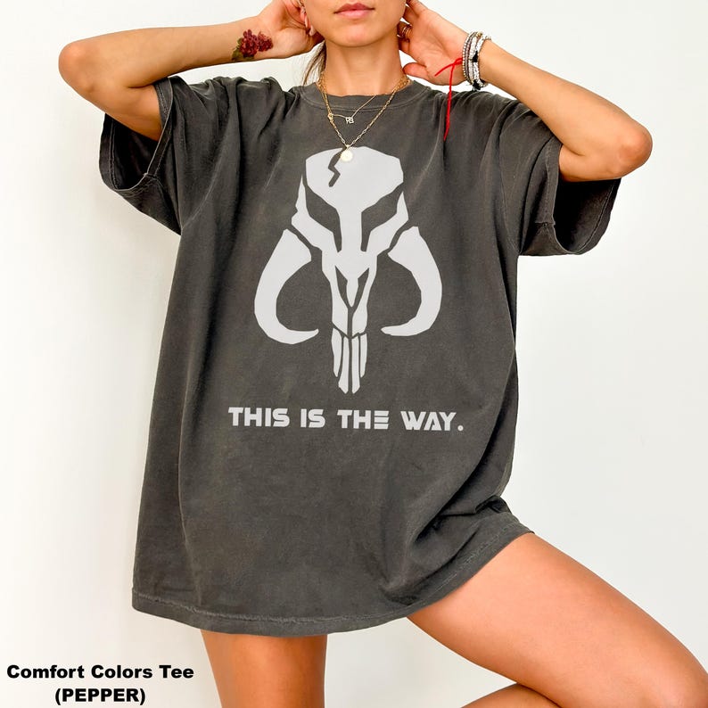 May include: Dark gray Comfort Colors tee featuring a white skull graphic with curved horns and the text "THIS IS THE WAY." The shirt is a relaxed fit, with short sleeves.